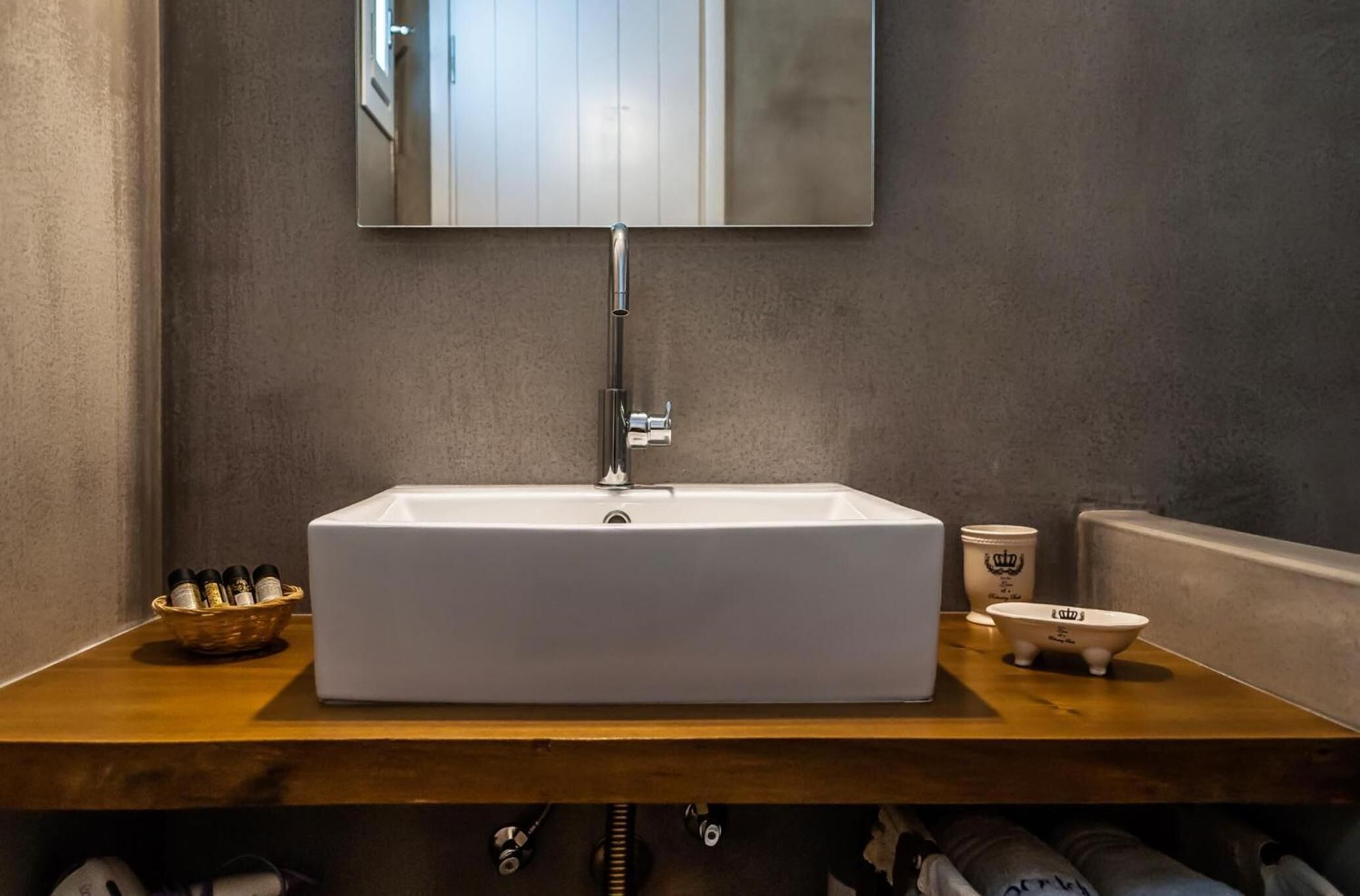 Un lavabo rectangulaire blanc, doté d’un robinet moderne, avec un miroir au-dessus, et un plan de travail en bois sur lequel se trouve un panier contenant des articles de toilette, ainsi qu’une petite baignoire blanche.