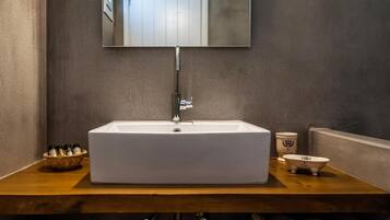 Un lavabo rectangulaire blanc, doté d’un robinet moderne, avec un miroir au-dessus, et un plan de travail en bois sur lequel se trouve un panier contenant des articles de toilette, ainsi qu’une petite baignoire blanche.