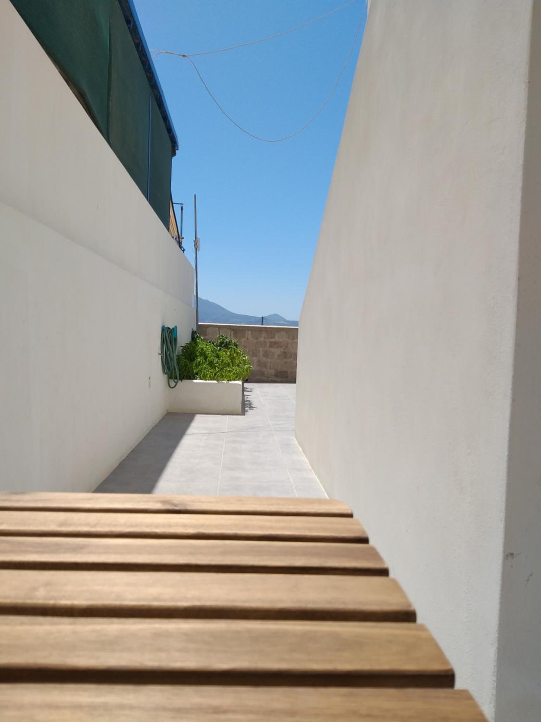 A narrow, sunlit corridor with a wooden bench at the end, leading to a white wall with a green awning.