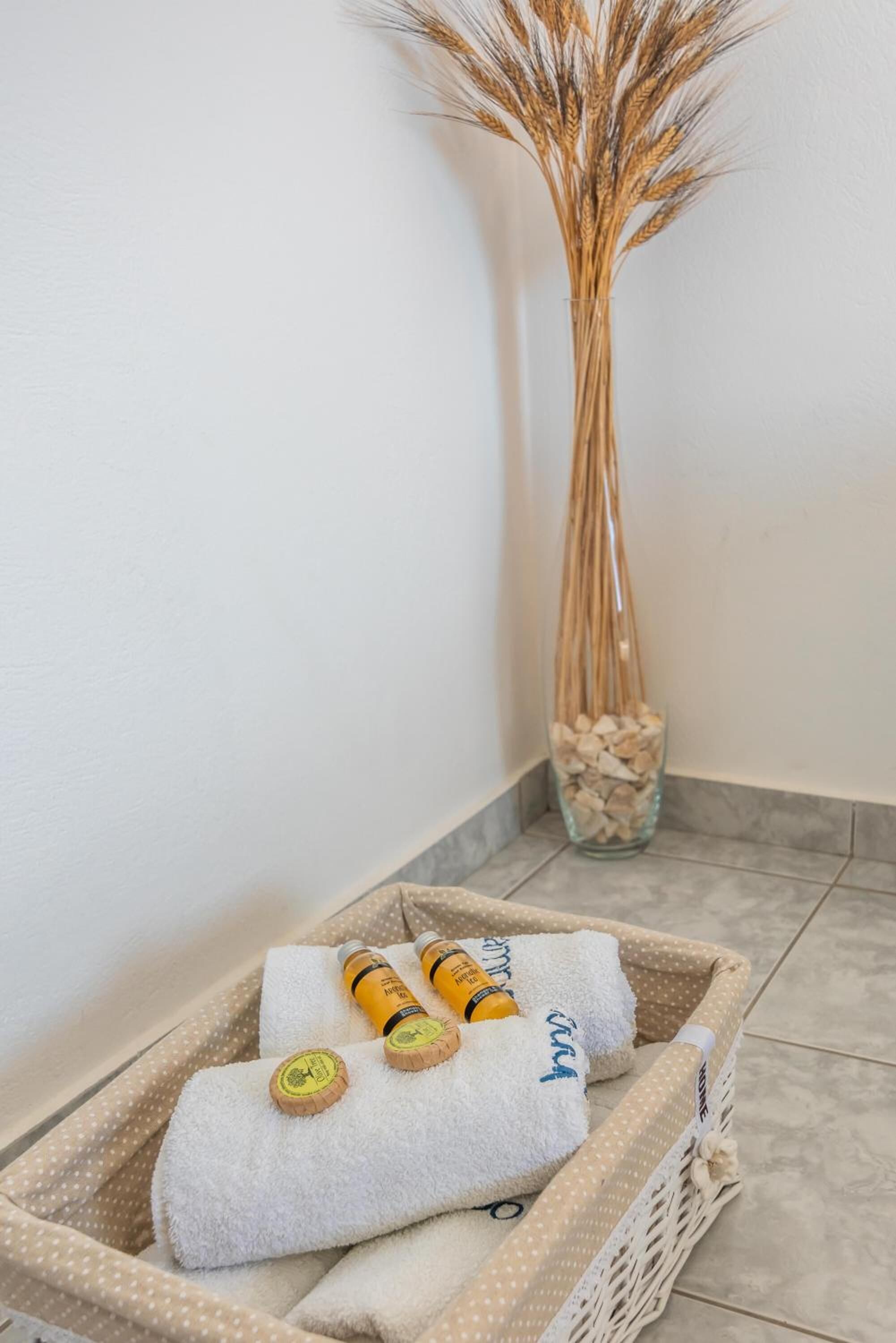 Esquina de una habitación con una pared blanca con textura, un jarrón de vidrio con plantas secas, y una cesta de mimbre que contiene una toalla blanca, dos botellas y un pequeño bloque de madera.
