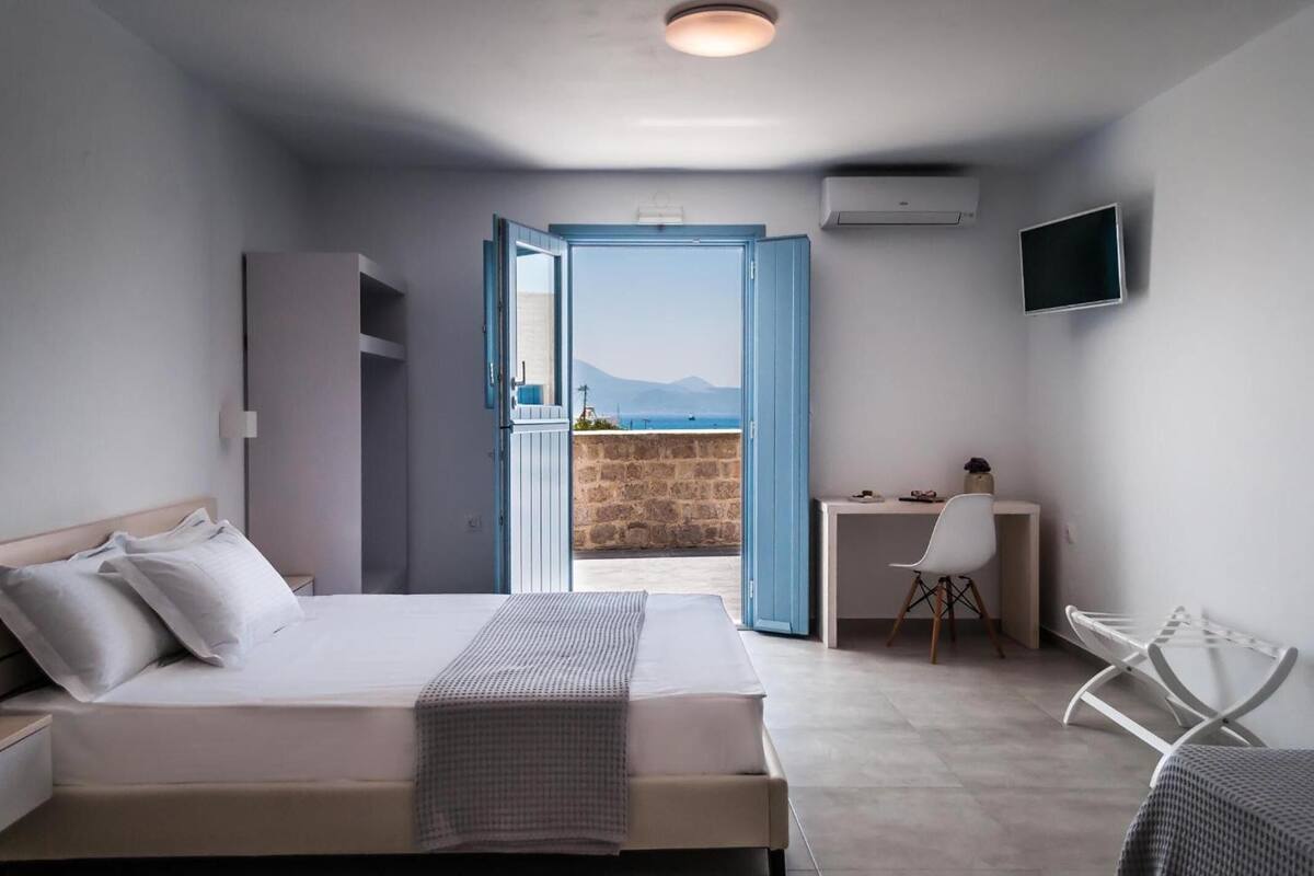 Quarto de hotel moderno com cama, escrivaninha e cadeira. Visão do mar e das montanhas através de uma porta aberta.