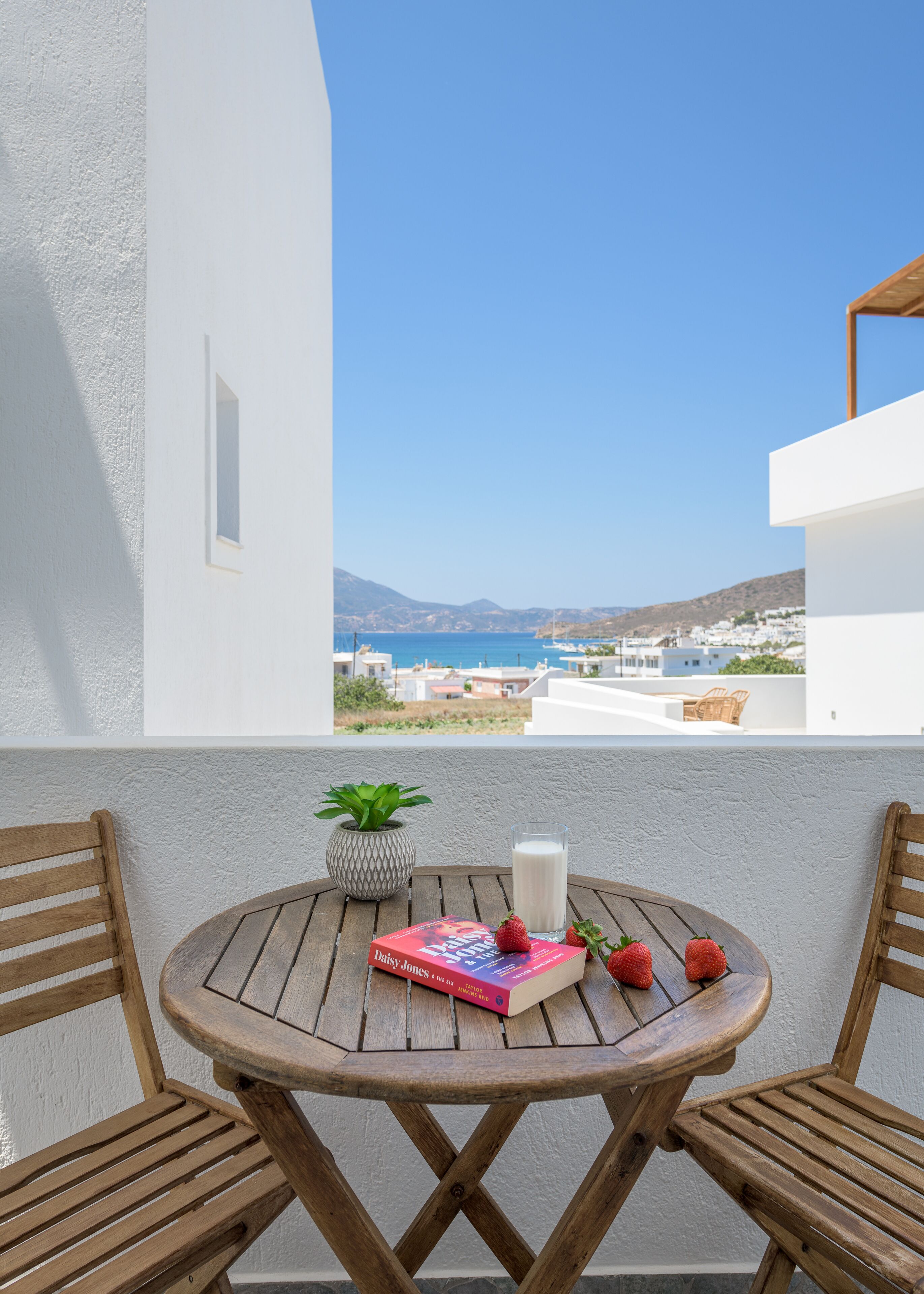 A wooden table with a book, a glass of milk, and strawberries on a balcony with a view of the sea and buildings.