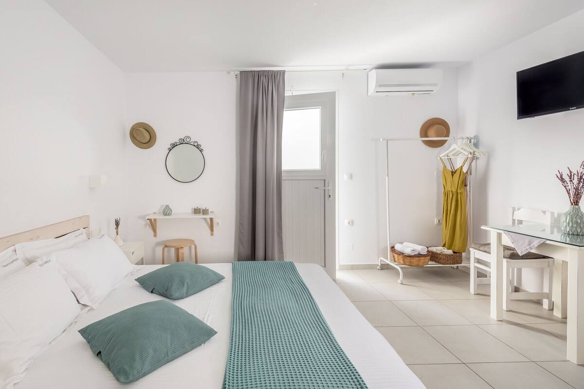 Quarto de hotel moderno com cama bem arrumada, uma pequena escrivaninha com cadeira, TV na parede e janela com cortinas.