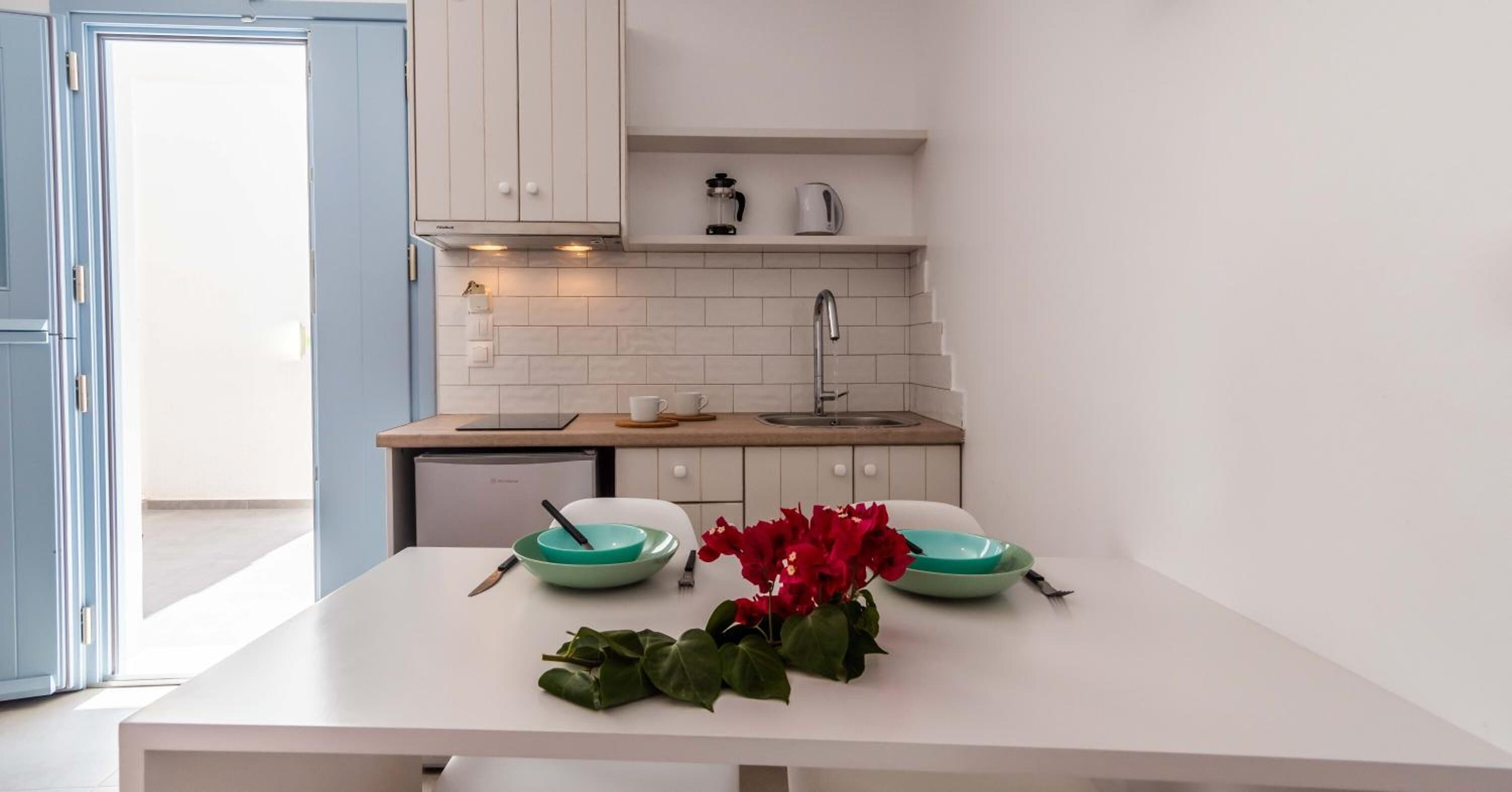 A modern kitchen with white cabinets, a white countertop, and a sink. There is a coffee maker on the countertop and a vase with red flowers on the table.