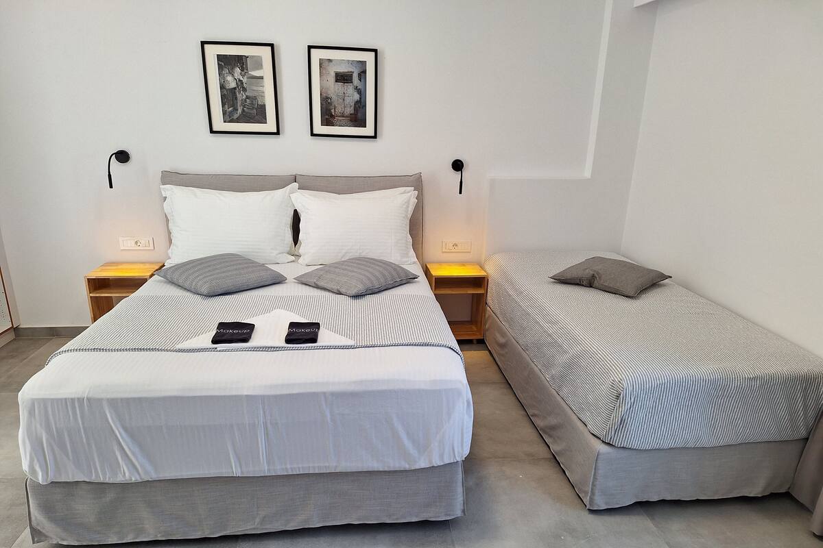 Uma cama de casal com cabeceira cinza e duas mesas de cabeceira com abajures, uma cama simples com colcha listrada, e duas fotografias emolduradas na parede.