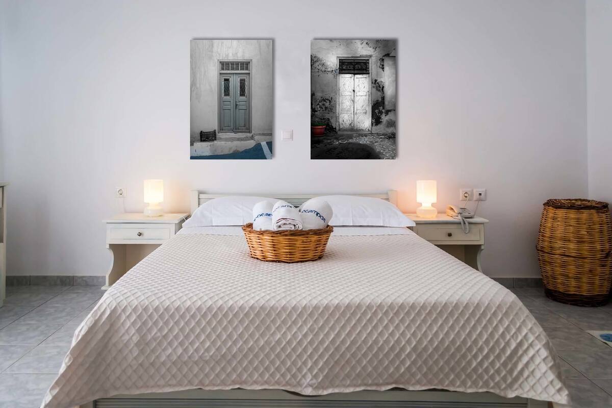 Quarto com cama, duas mesas de cabeceira, cesta de toalhas e duas luminárias fixadas na parede.