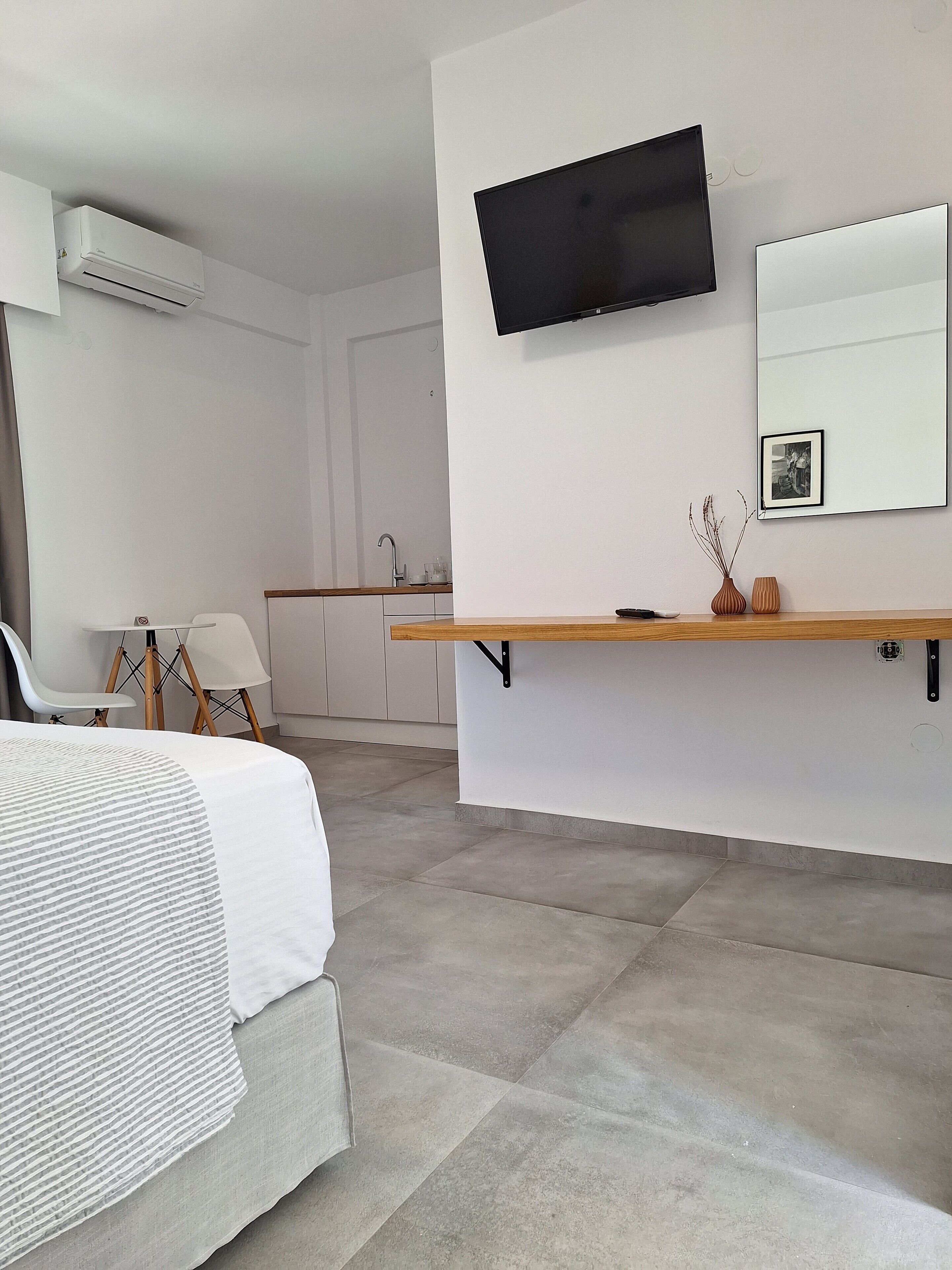 A modern hotel room with a bed, a TV mounted on the wall, a small kitchen area, and a wooden shelf with decorative items.