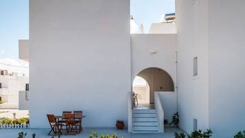 A white building with a staircase leading to an upper level. There is a table and chairs outside on the ground floor.
