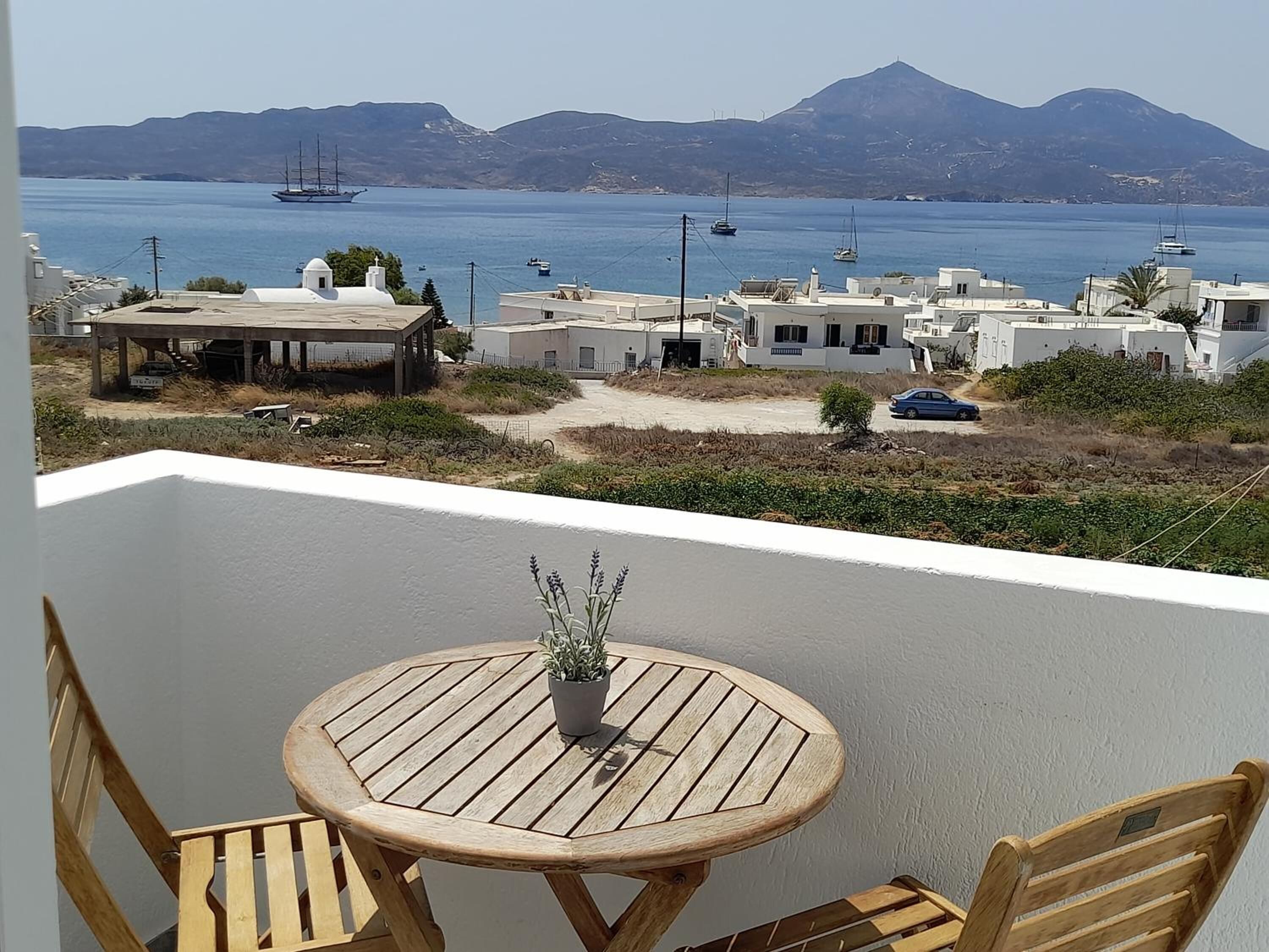 Un balcon doté d’une table ronde en bois et de chaises, offrant une vue sur une zone côtière avec des bâtiments et des bateaux.