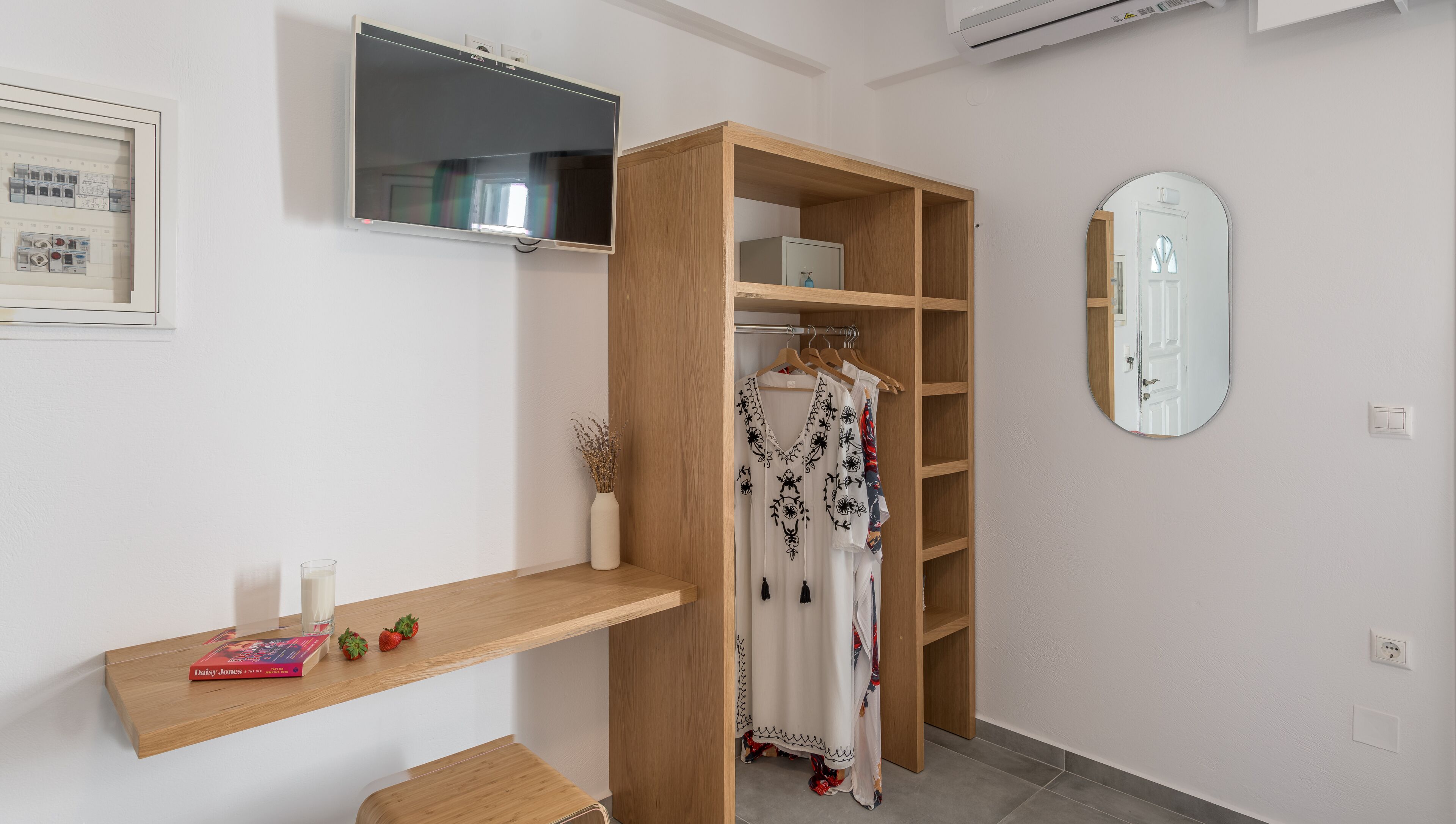 Een kamer met een houten garderobe, een wandgemonteerde televisie, een spiegel en een plank met boeken en aardbeien.