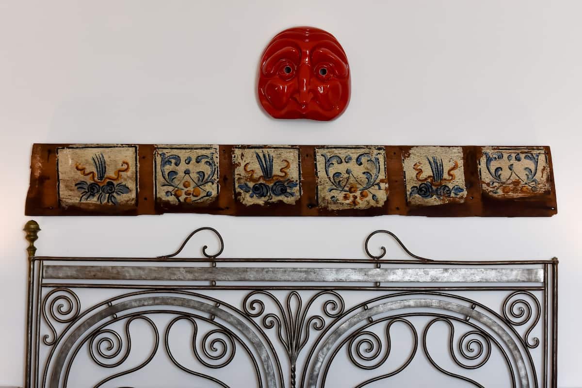 A wall-mounted decorative piece with a red face, a metal headboard with intricate scrollwork, and a series of tiles with floral patterns.