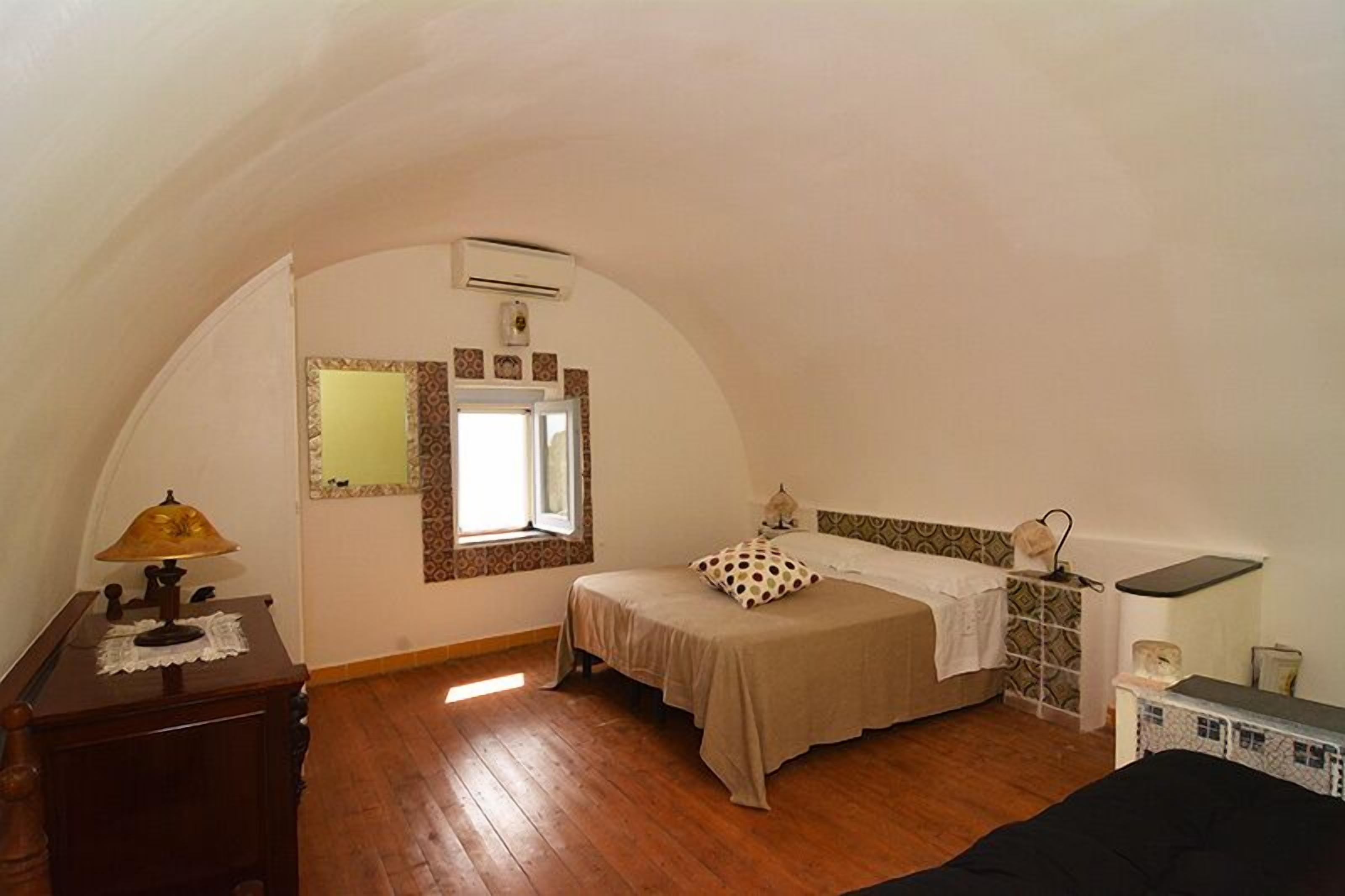 A small, cozy bedroom with a single bed, a bedside table with a lamp, a window with patterned curtains, and a small shelf with a decorative item.