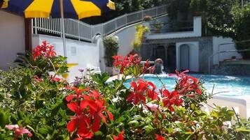 Area piscina con fiori rossi in primo piano, un ombrellone giallo e blu, e un edificio bianco con una scala sullo sfondo.