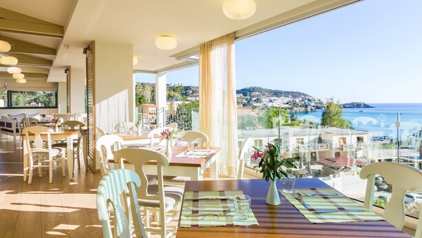 En spiseplads med borde dækket til to, store vinduer med udsigt over havet og byen, samt en balkon med udendørs siddepladser.