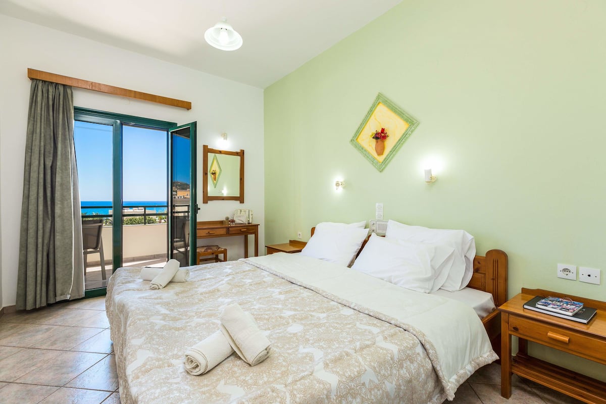 A hotel room with a bed, a desk, a chair, a nightstand, a window with a view, and a wall-mounted light fixture.