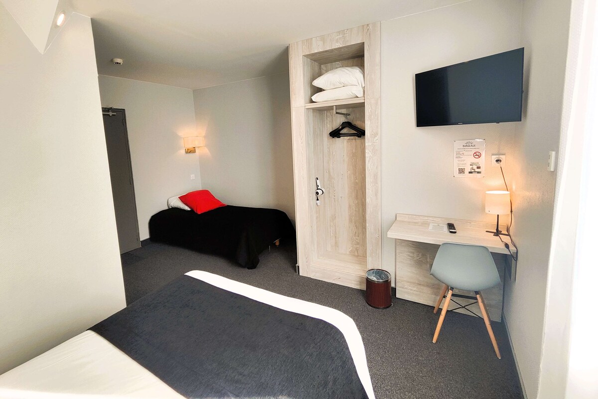 Habitación de hotel compacta con una cama individual, un escritorio con silla, un televisor montado en la pared y un armario con perchero.
