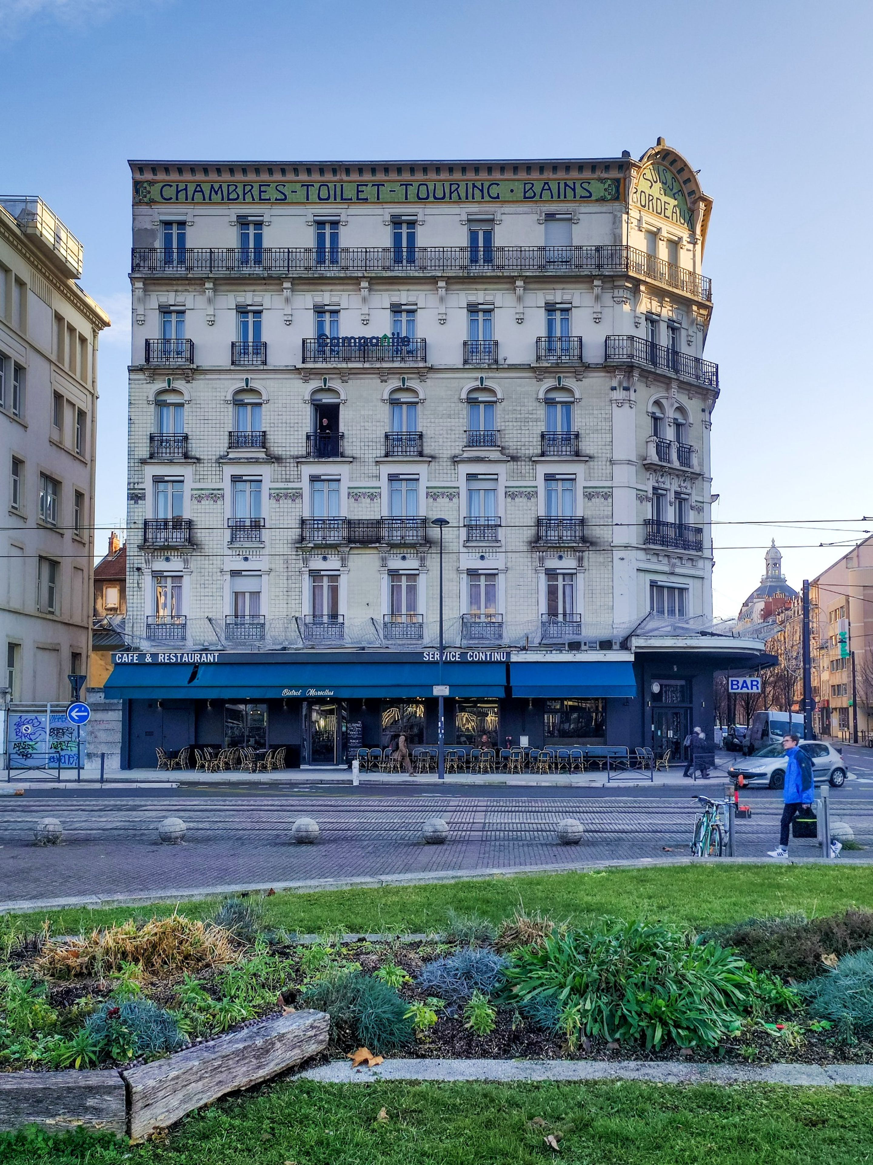 Un immeuble à plusieurs étages abritant un café et un restaurant au rez-de-chaussée, une enseigne indiquant la continuité des services et un bar.