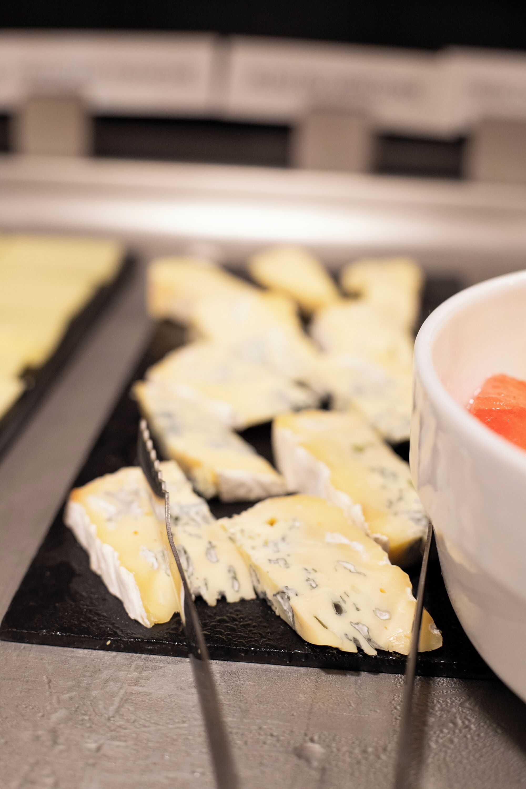 A close-up of a cheese board with various types of cheese and a bowl of fruit.