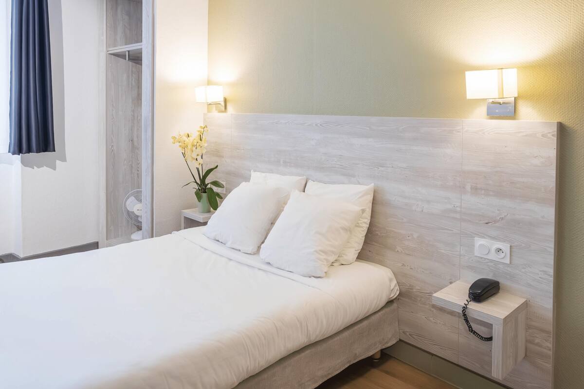 A neatly made bed with white linens, a wooden headboard, bedside lamps, a small bedside table with a phone, and a potted plant.