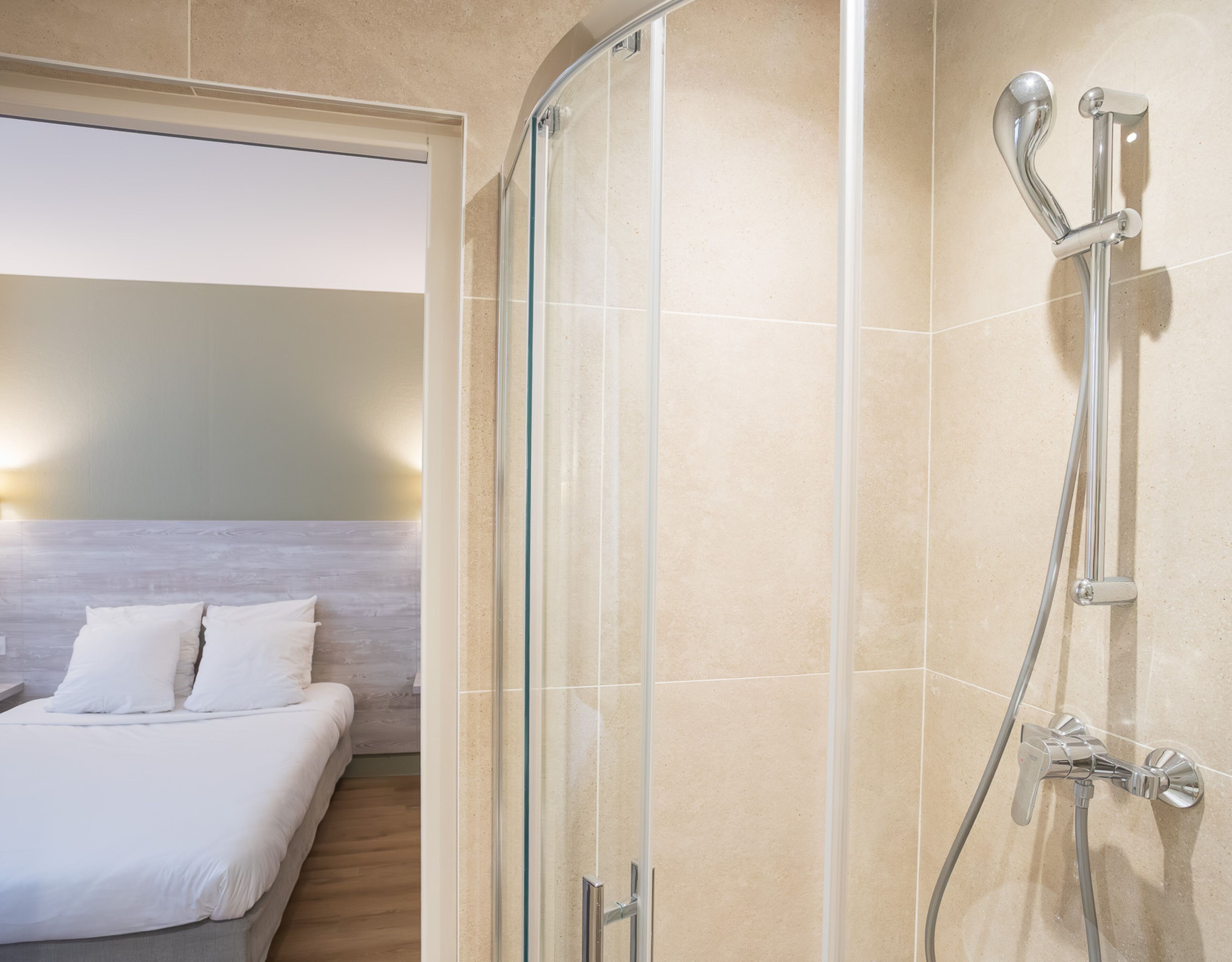 A hotel room with a glass-enclosed shower, a bed with white bedding, and a wooden headboard.