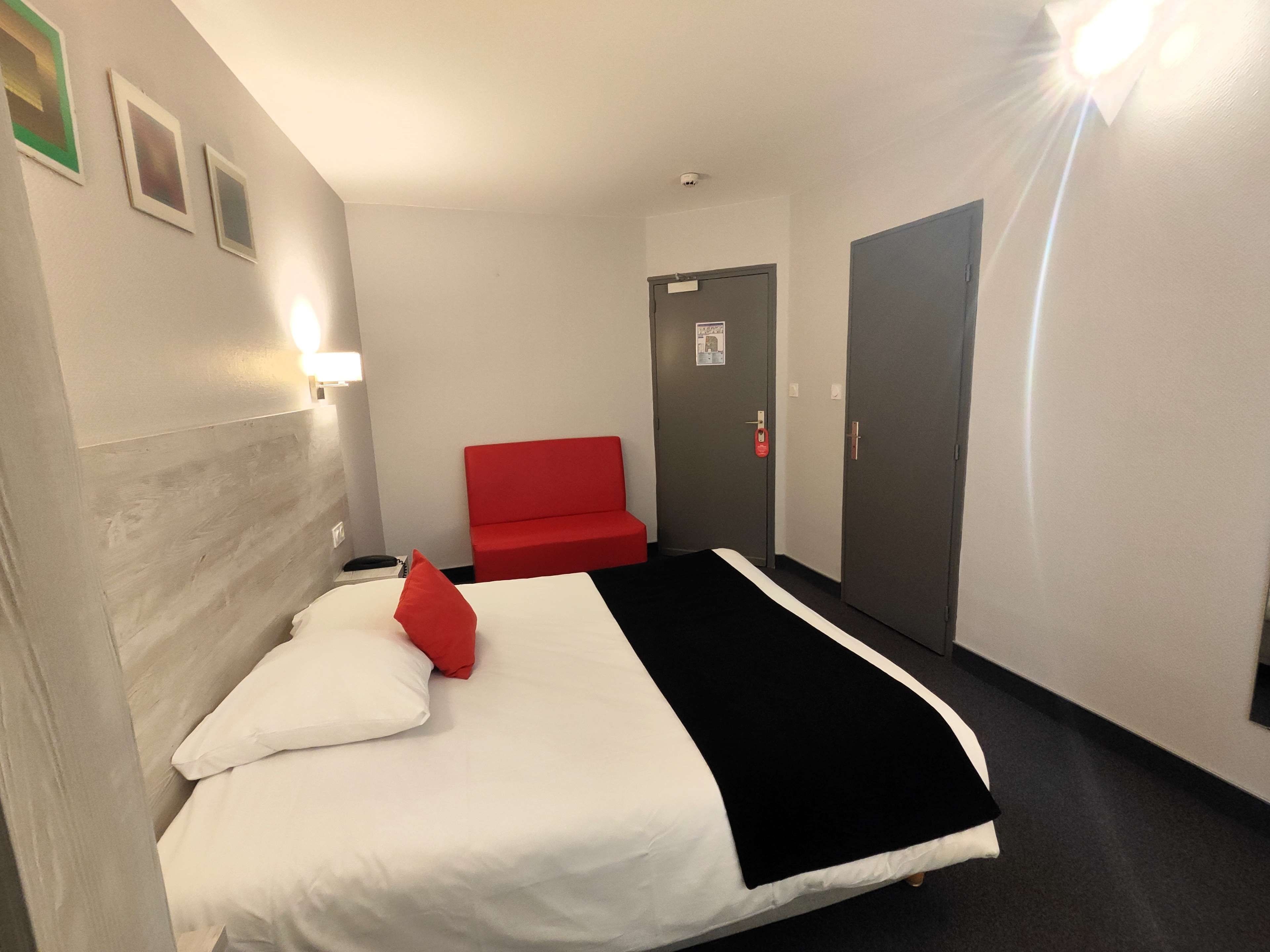 Habitación de hotel con cama, una silla roja, una puerta y una lámpara fijada a la pared.