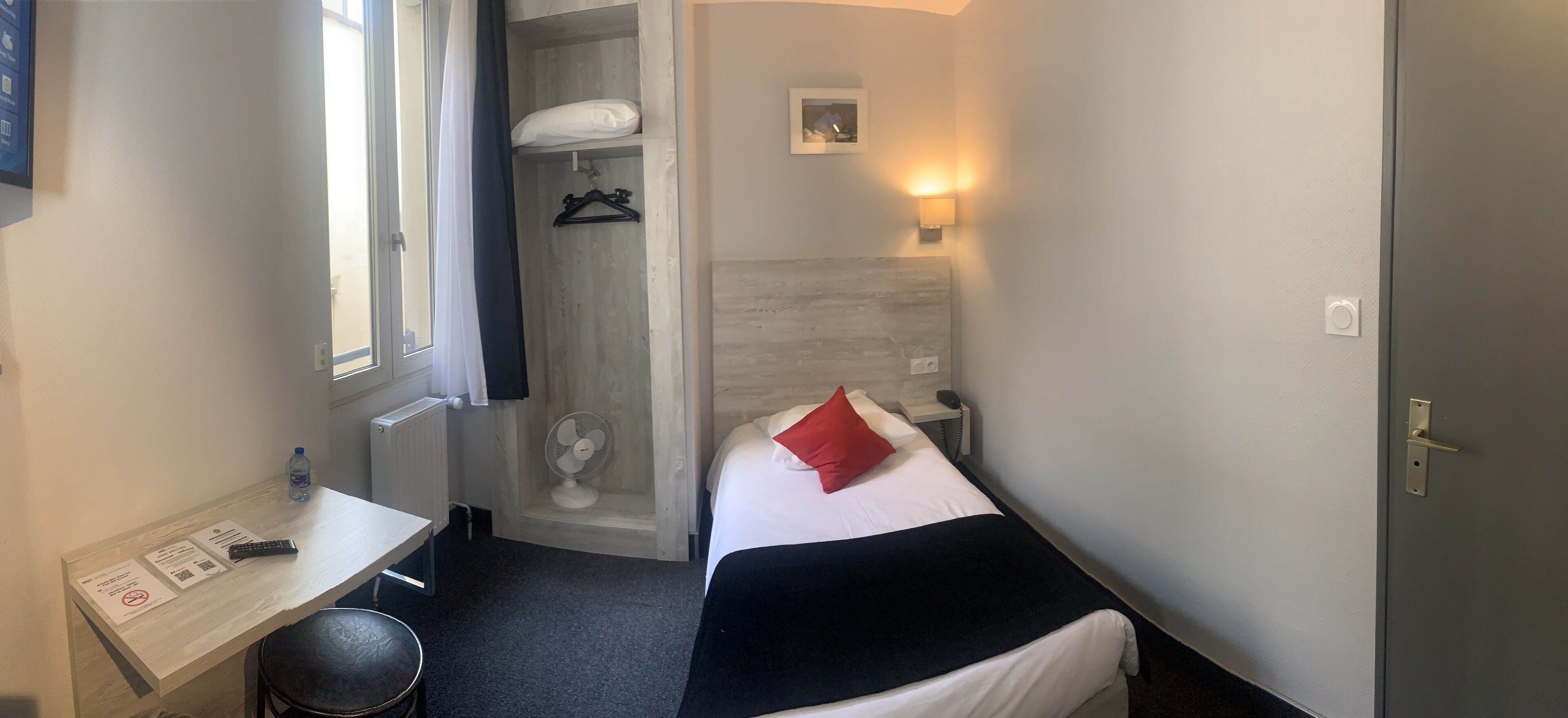 Ett kompakt hotellrum med en enkel säng, ett nattduksbord med en fjärrkontroll, ett litet skrivbord med en stol, en väggmonterad fläkt och ett fönster med en gardin.
