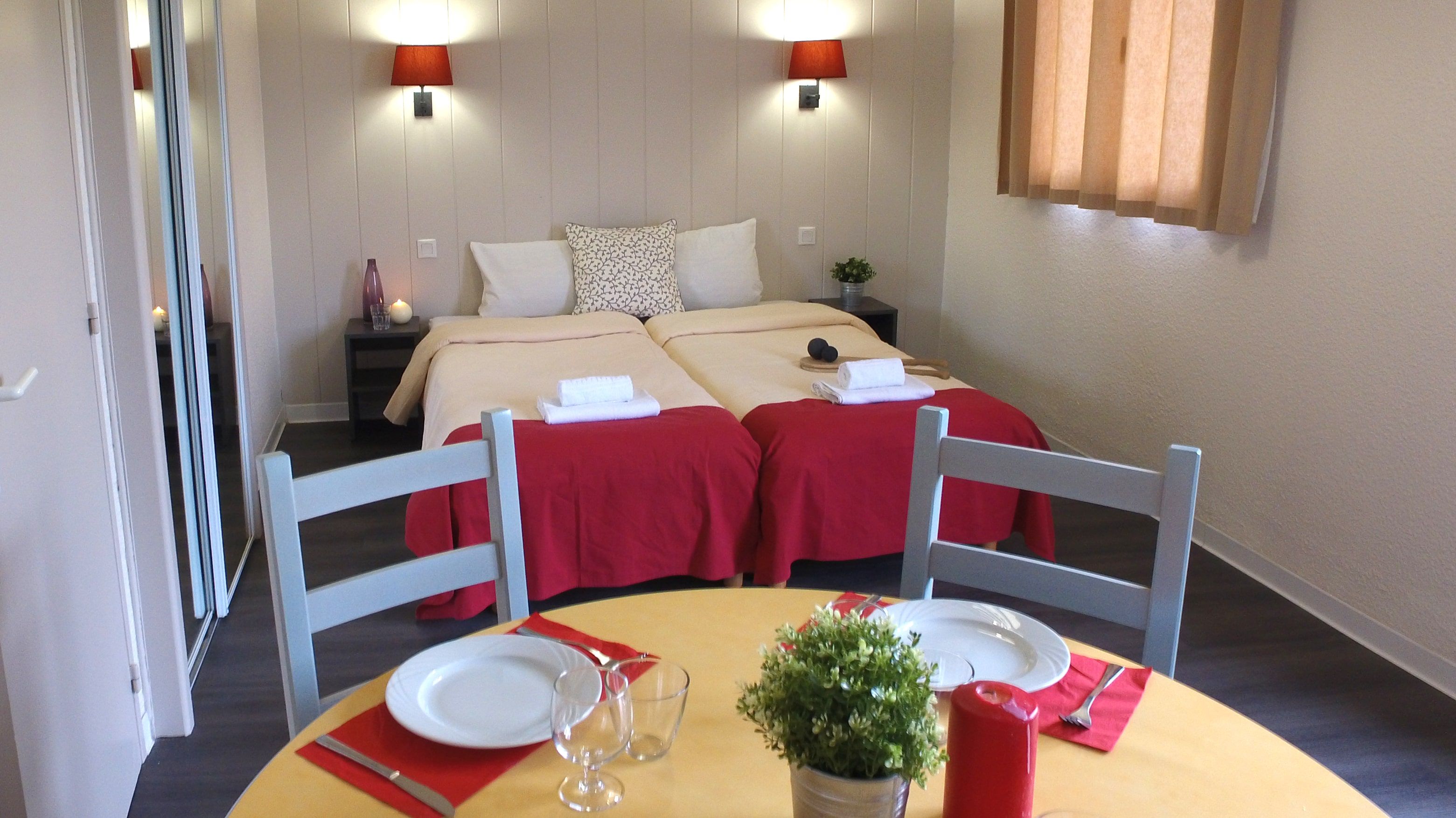 Una habitación de hotel con una cama, dos mesitas de noche, una pequeña mesa con una planta y un rincón de comedor con sillas y una mesa preparada para dos.