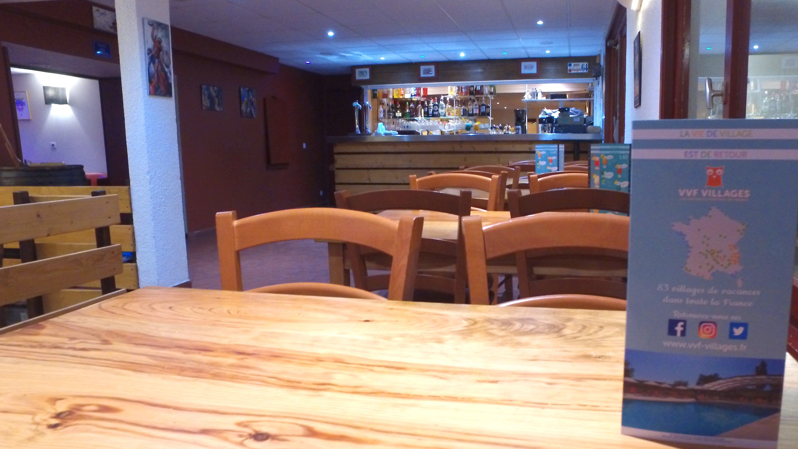 Een restaurantinterieur met houten tafels en stoelen, een bar op de achtergrond, en een bord met informatie.