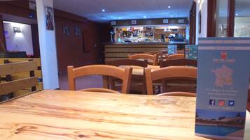 Een restaurantinterieur met houten tafels en stoelen, een bar op de achtergrond, en een bord met informatie.