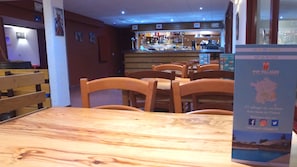 Interior de un restaurante con mesas y sillas de madera, una zona de bar al fondo y un cartel con información.