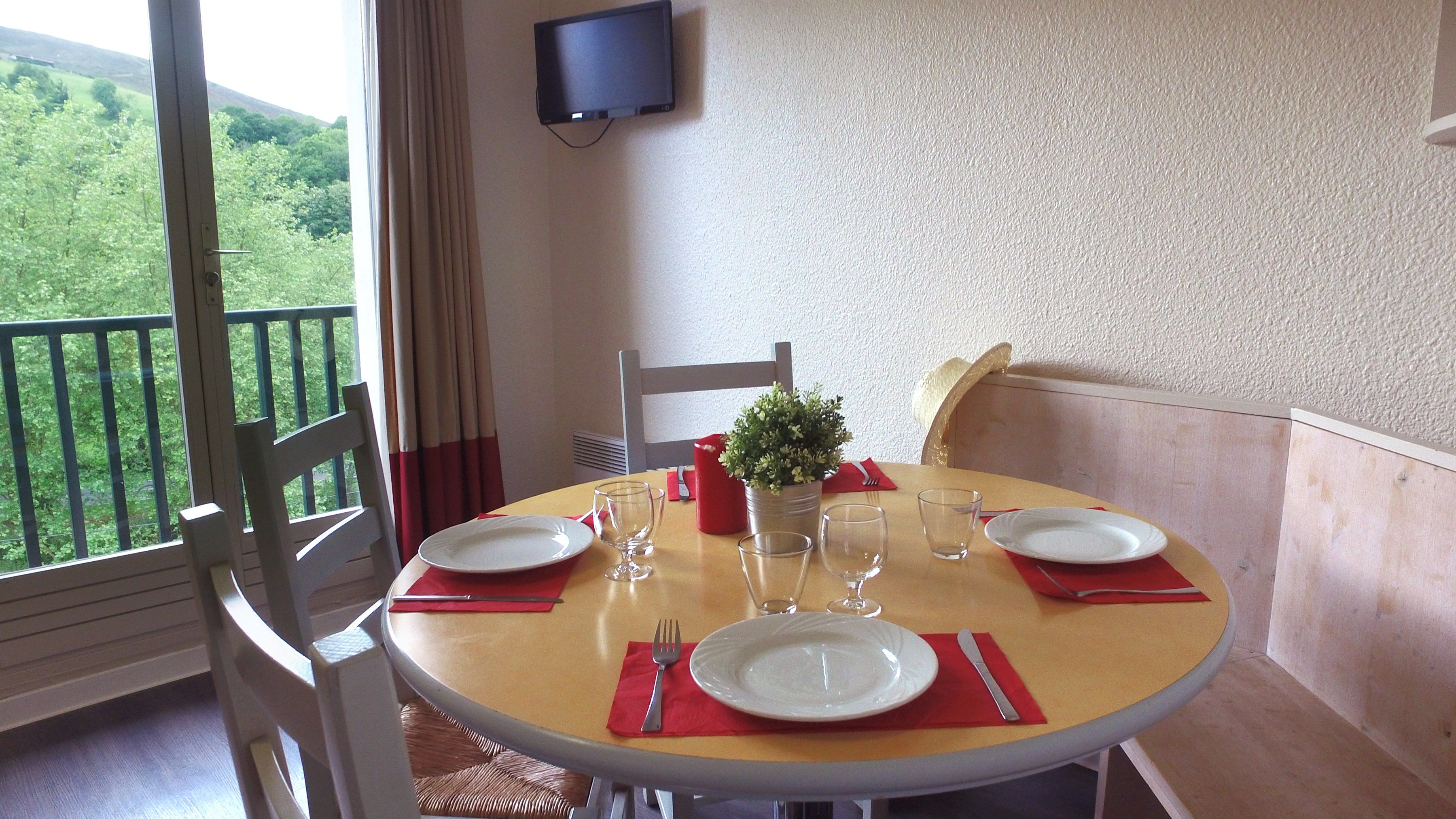 A dining table set for two with plates, cutlery, and glasses, a small potted plant, and a red candle on a balcony overlooking greenery.