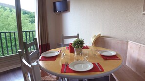 Mesa de comedor lista para dos personas con platos, cubiertos y vasos, una pequeña planta en maceta y una vela roja en un balcón con vistas a la vegetación.