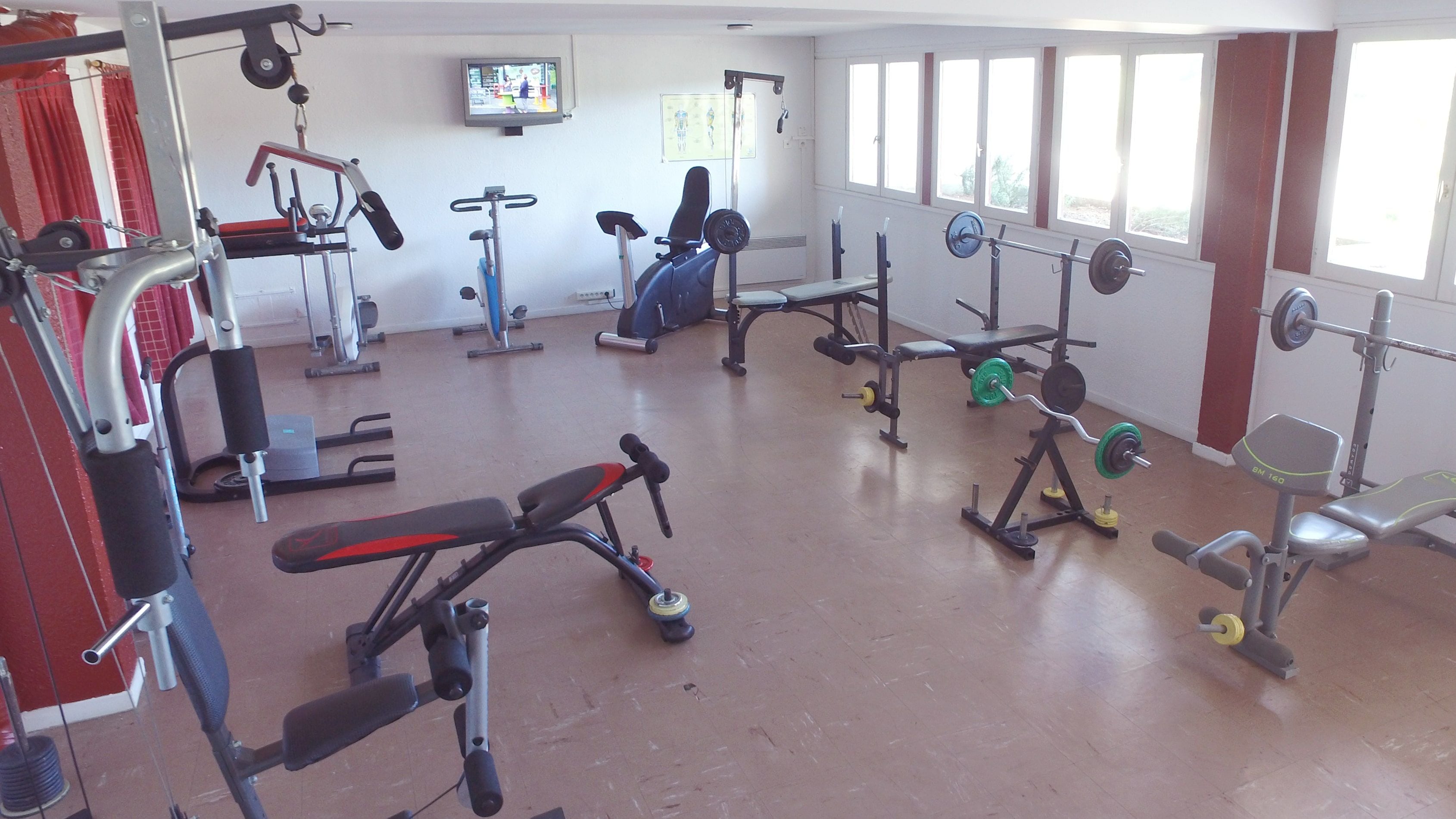 Une salle de sport équipée de divers appareils d’exercice, avec un téléviseur fixé au mur et de grandes fenêtres.