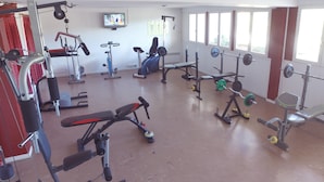 Un gimnasio con diversos equipos de ejercicio, un televisor en la pared y ventanas amplias.