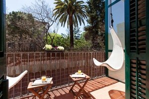 A balcony with two wooden tables, a hanging chair, and a view of trees and a palm tree. - Hostal Ecozentric (Barcelona)
