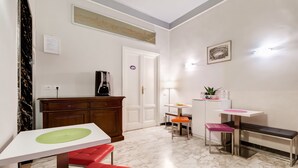 A compact hotel room with a white marble floor, a dining area with a white table and red bench, a small kitchenette, and a door leading to another room. - B&B 2 Passi Al Colosseo (Rome)