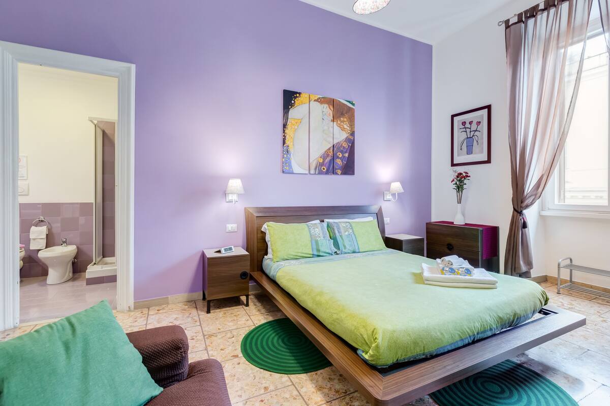 A modern bedroom with a bed, nightstands, a painting, and a bathroom visible in the background.