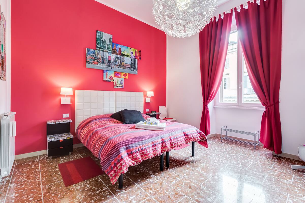 A bedroom with a red wall, a white headboard, a bed with a patterned bedspread, two bedside tables with lamps, and a window with red curtains.