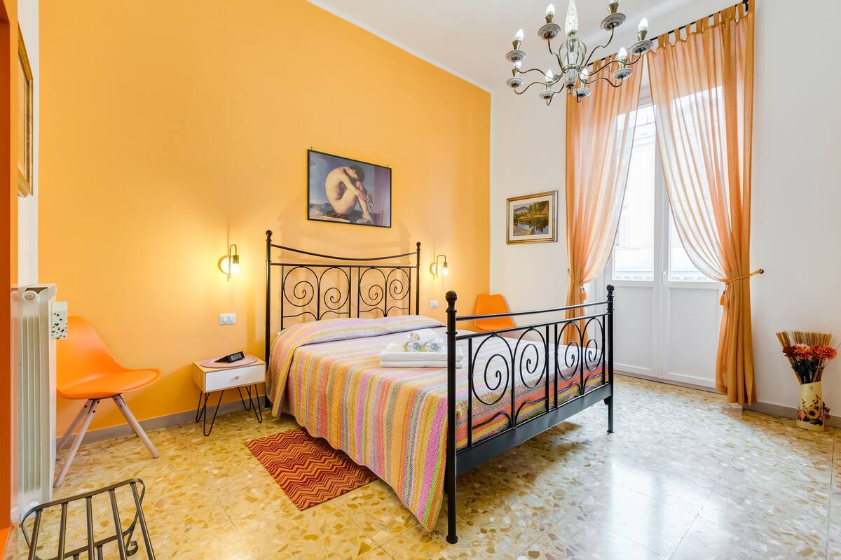 A bedroom with a bed, a chair, a radiator, a nightstand, and a window with curtains.