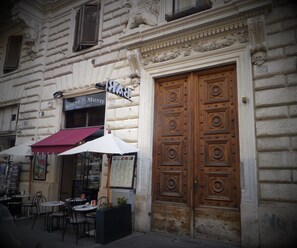 A building with a large wooden door and a sign above it. There are outdoor tables and chairs in front of the building. - B&B 2 Passi Al Colosseo (Rome)