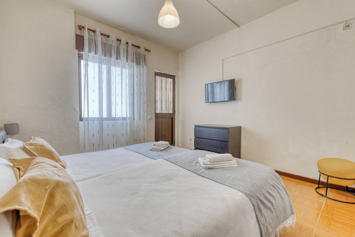 A bedroom with a bed, a desk, a chair, a TV, and a window with curtains.