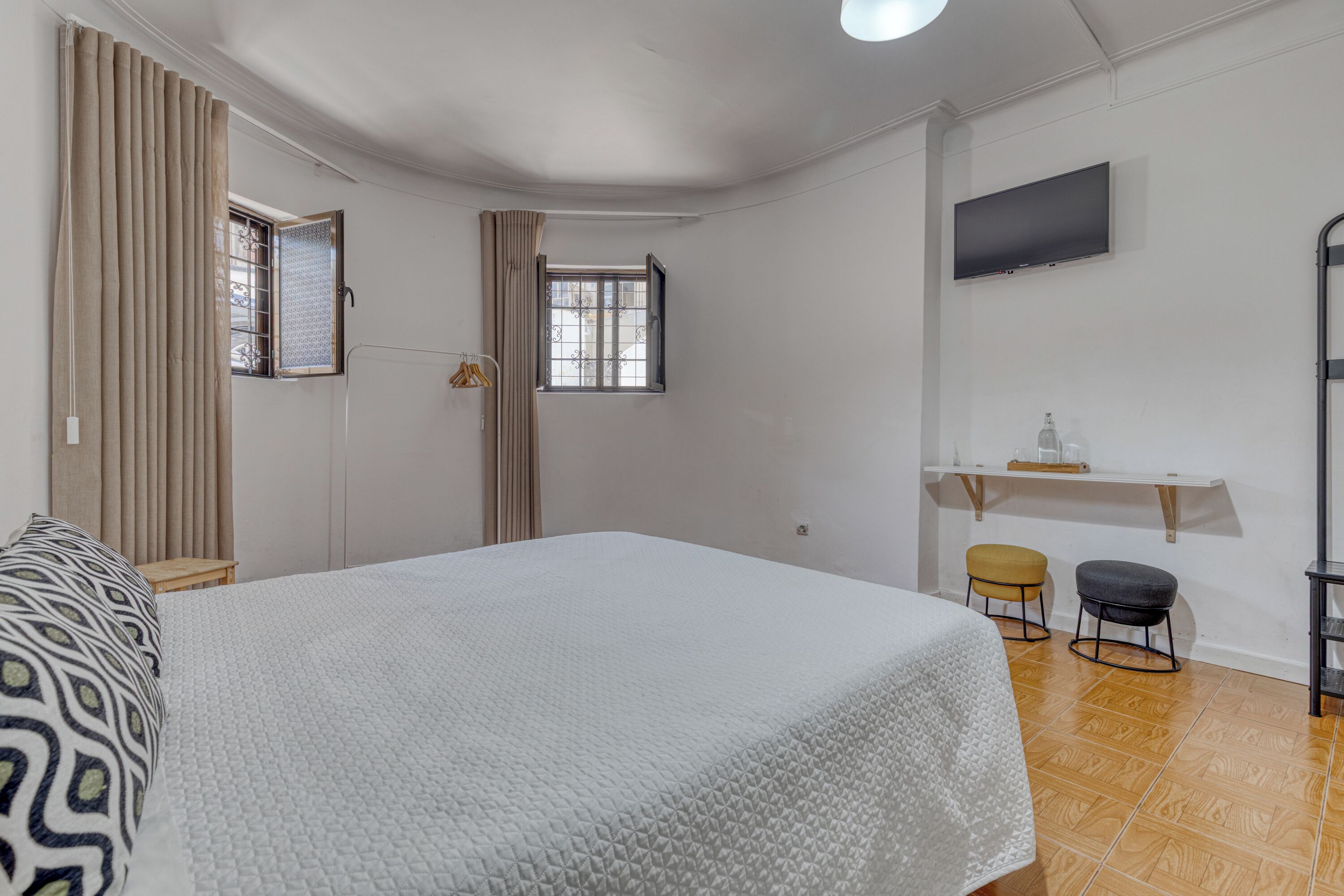 A bedroom with a large bed, a TV mounted on the wall, a small table with a chair, and a window with curtains.