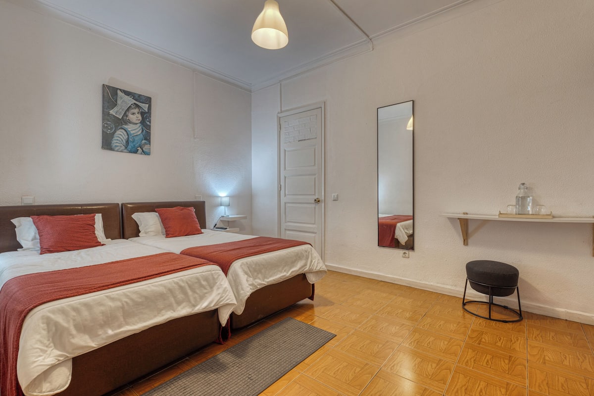 A hotel room with two beds, a wooden floor, a small stool, a nightstand, a mirror, and a painting on the wall.