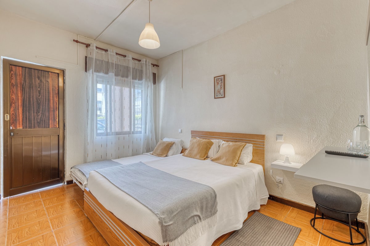 A bedroom with a wooden door, a bed with a grey blanket, a bedside table with a lamp, and a small stool.
