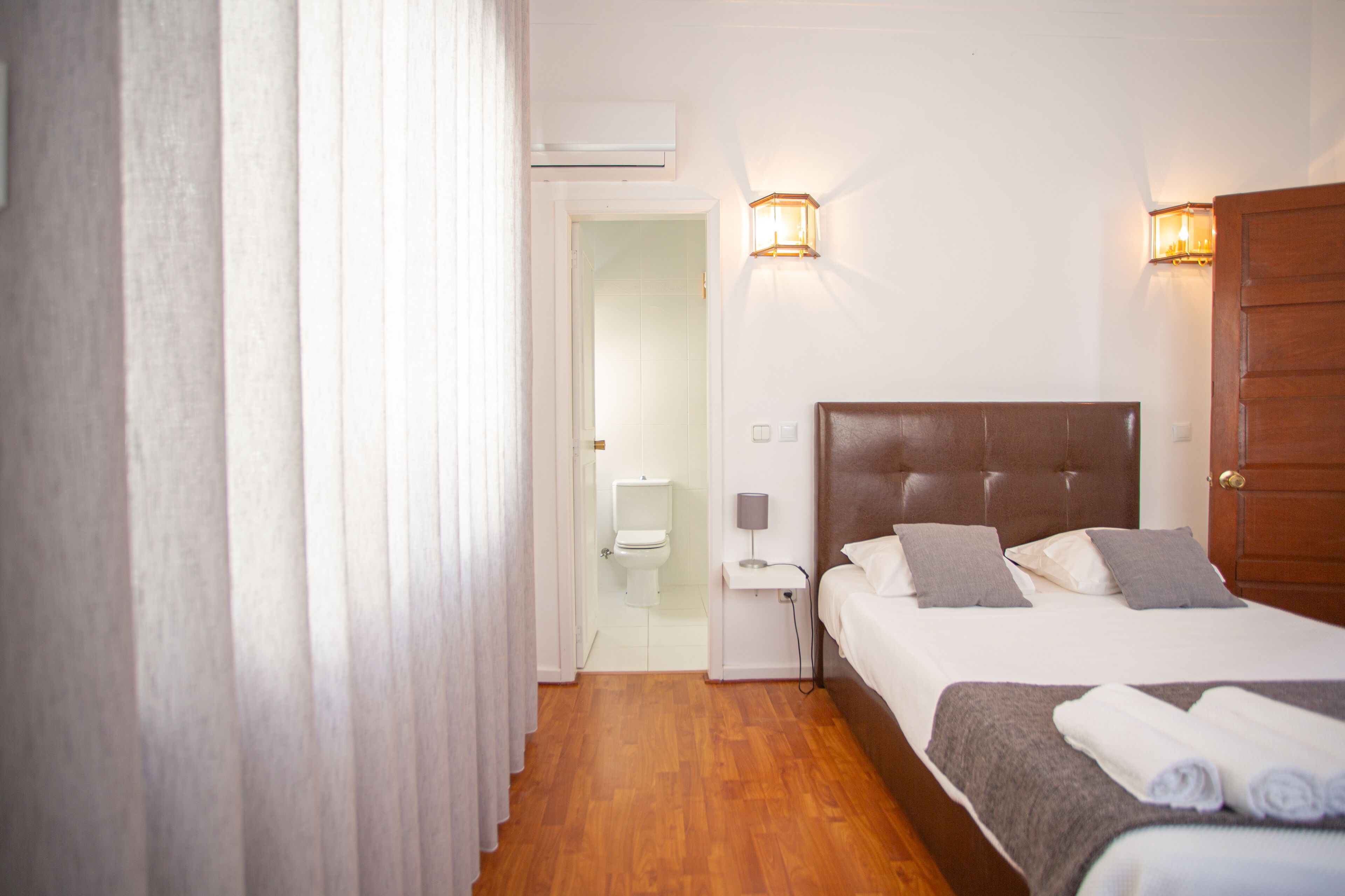 A hotel room with a wooden floor, a bed with a headboard, a nightstand, a lamp, and a wall-mounted light fixture.