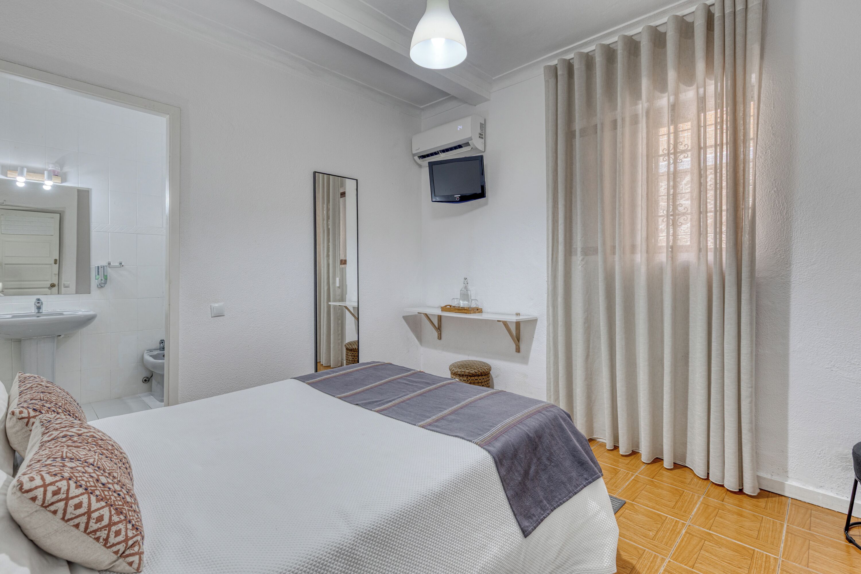 A bedroom with a bed, a bedside table, a mirror, a TV, a shelf, and a window with curtains.
