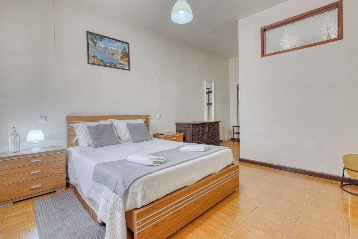 A bedroom with a wooden bed, a nightstand, a dresser, a painting, and a window.