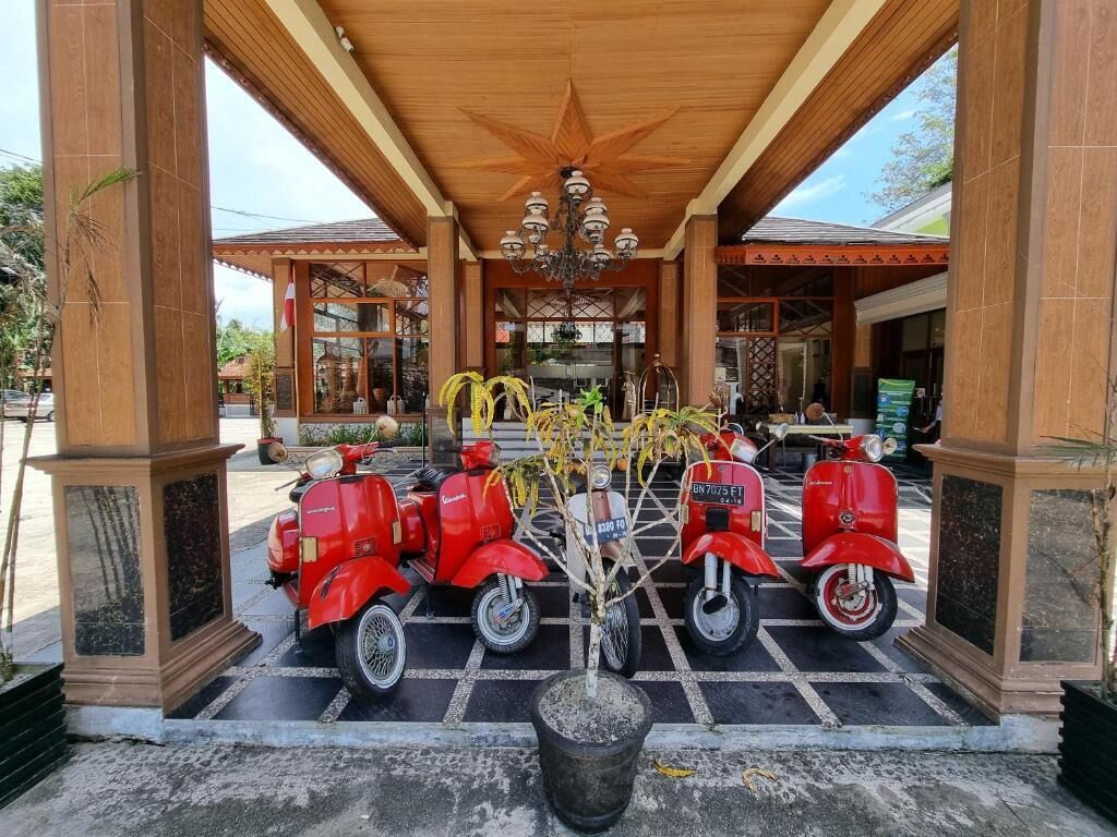 Un ingresso coperto con soffitto in legno e lampadario, tre scooter rossi parcheggiati davanti e una pianta in vaso al centro.