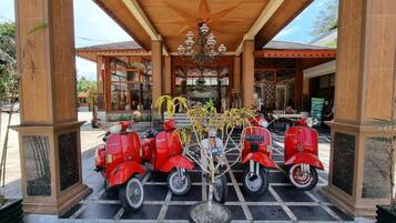 Un ingresso coperto con soffitto in legno e lampadario, tre scooter rossi parcheggiati davanti e una pianta in vaso al centro.