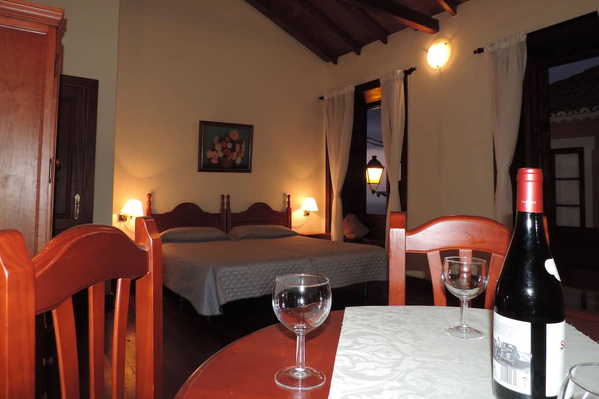 Una habitación de hotel con una cama, dos mesitas de noche, una mesa con una botella de vino y copas, y un cuadro en la pared.
